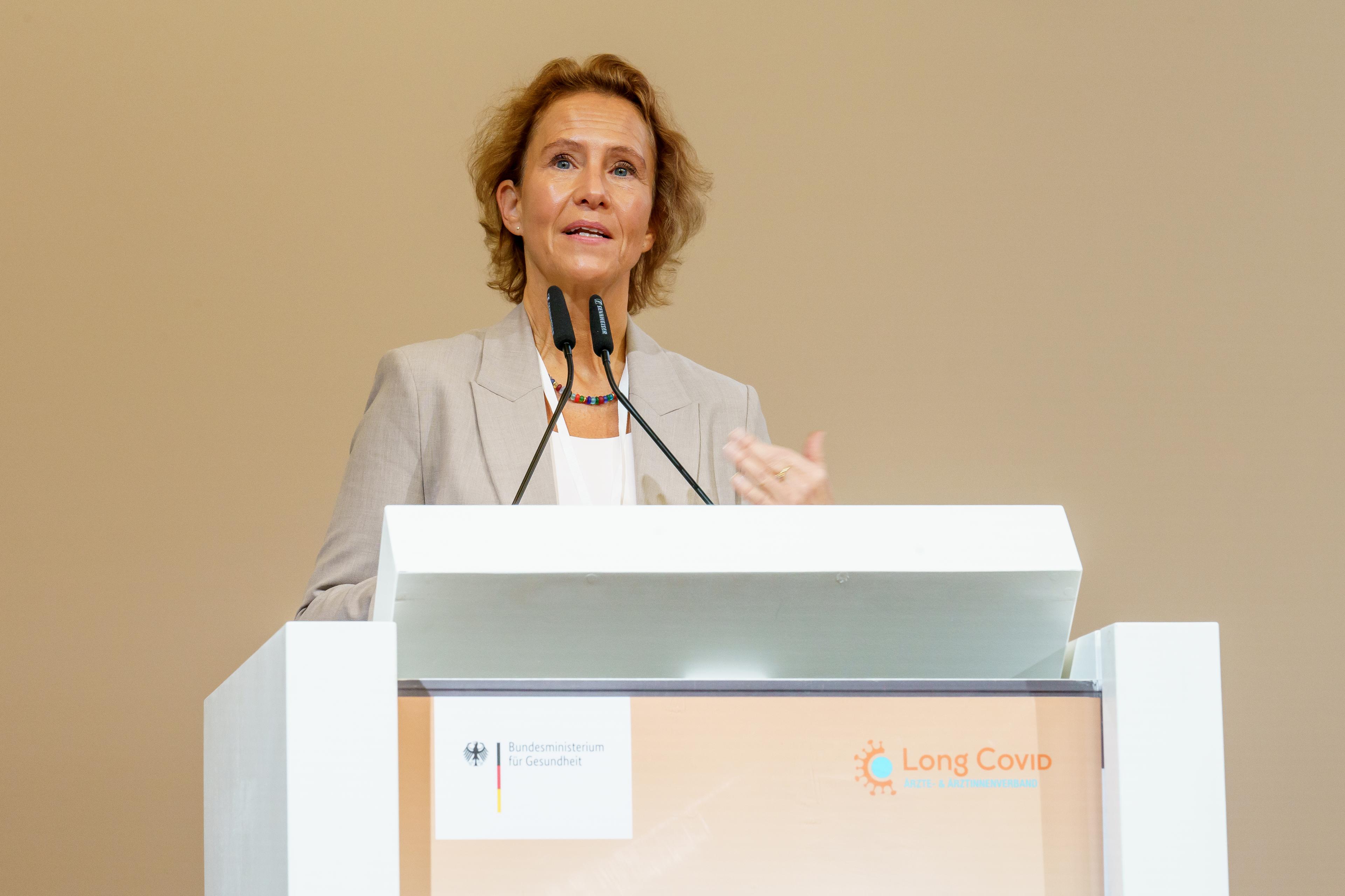 Prof. Dr. Christine Falk steht am Rednerpult und hält eine Rede auf dem 3. Long-COVID-Kongress in Berlin. Sie trägt einen hellen Blazer und spricht vor zwei Mikrofonen. Das Rednerpult ist mit Logos des Bundesministeriums für Gesundheit und des Ärzte- und Ärztinnenverbands Long COVID versehen.