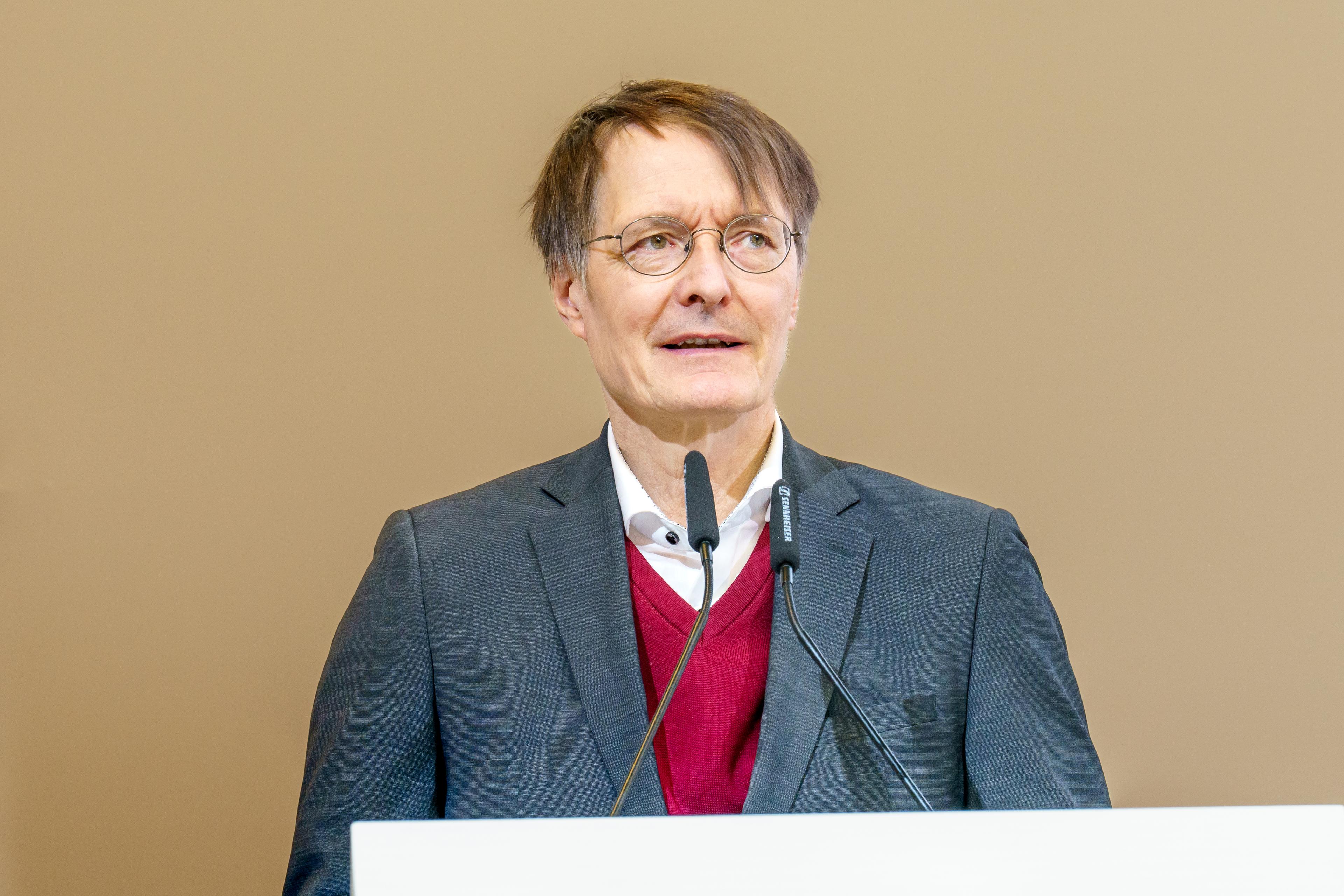 Prof. Dr. Karl Lauterbach, Bundesgesundheitsminister, steht am Rednerpult und spricht auf dem 3. Long-COVID-Kongress in Berlin. Er trägt einen grauen Anzug, ein weißes Hemd und einen roten Pullover unter dem Sakko. Zwei Mikrofone sind vor ihm angebracht.